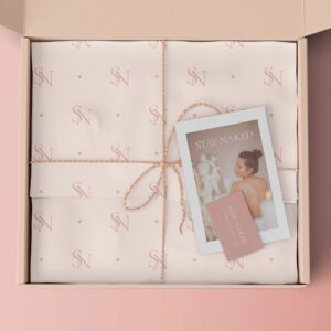 An open box displaying neatly wrapped items in cream-colored paper with a pink SN logo pattern tied with twine A Stay Naked card featuring a woman with a towel is placed on top