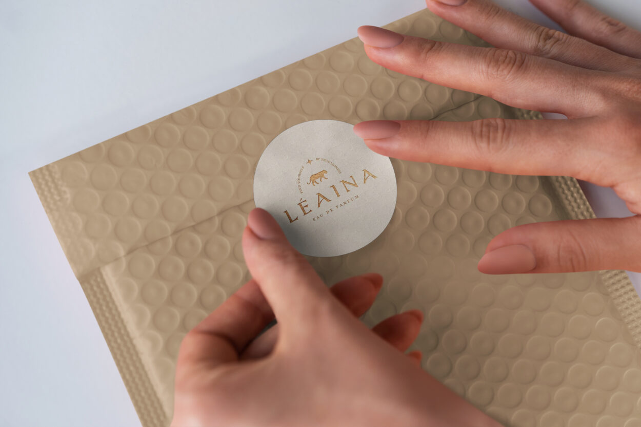 Close-up of hands sealing a beige bubble mailer with a round white sticker featuring the brand 'Léaina Eau de Parfum' in gold lettering.