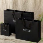 Three black shopping bags of varying sizes with the "NOTRE" logo in white, placed against a beige fabric backdrop.