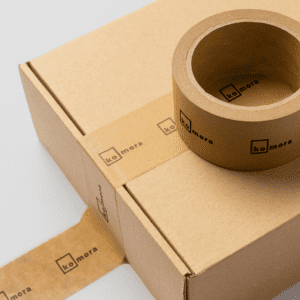 A cardboard box sealed with brown kraft tape featuring the "komora" logo, with an additional roll of the same branded tape placed on top.