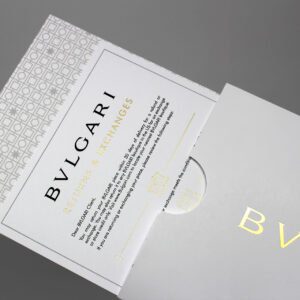 Bulgari returns and exchanges card partially visible inside an elegant white envelope with gold embossed "Bulgari" logo, featuring a decorative gray pattern on the card.