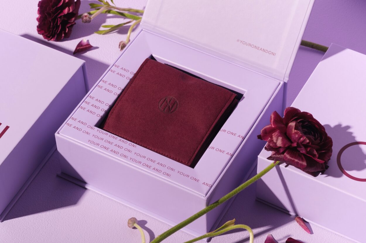 A close-up of an open white gift box with the phrase 'Your One and Only' printed on its inner edges. Inside, a burgundy fabric pouch is embossed with a circular monogram logo. The box is accompanied by a deep burgundy flower, placed artistically nearby, adding a touch of elegance and sophistication.