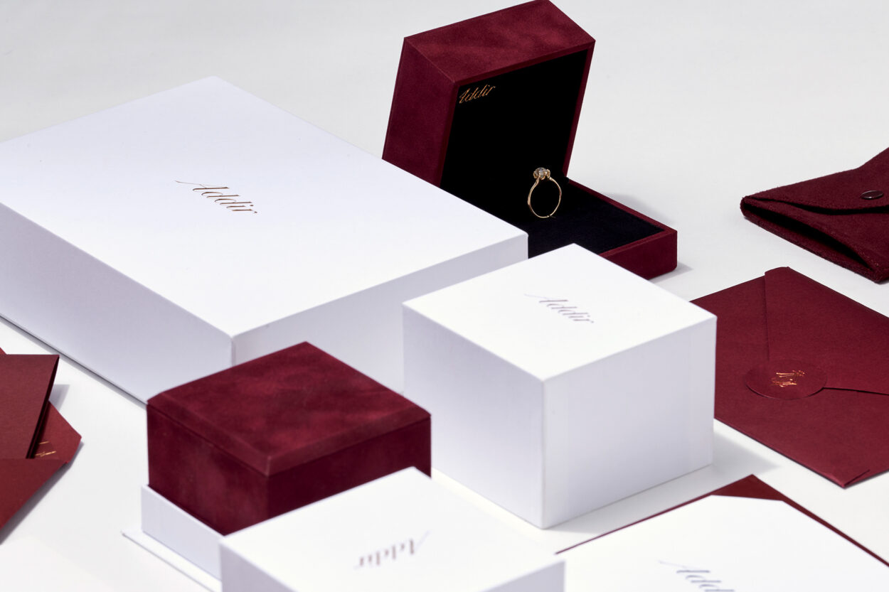 A collection of Adair branded packaging boxes in white and burgundy velvet along with envelopes, showcasing a ring in an open burgundy velvet box on a white surface.