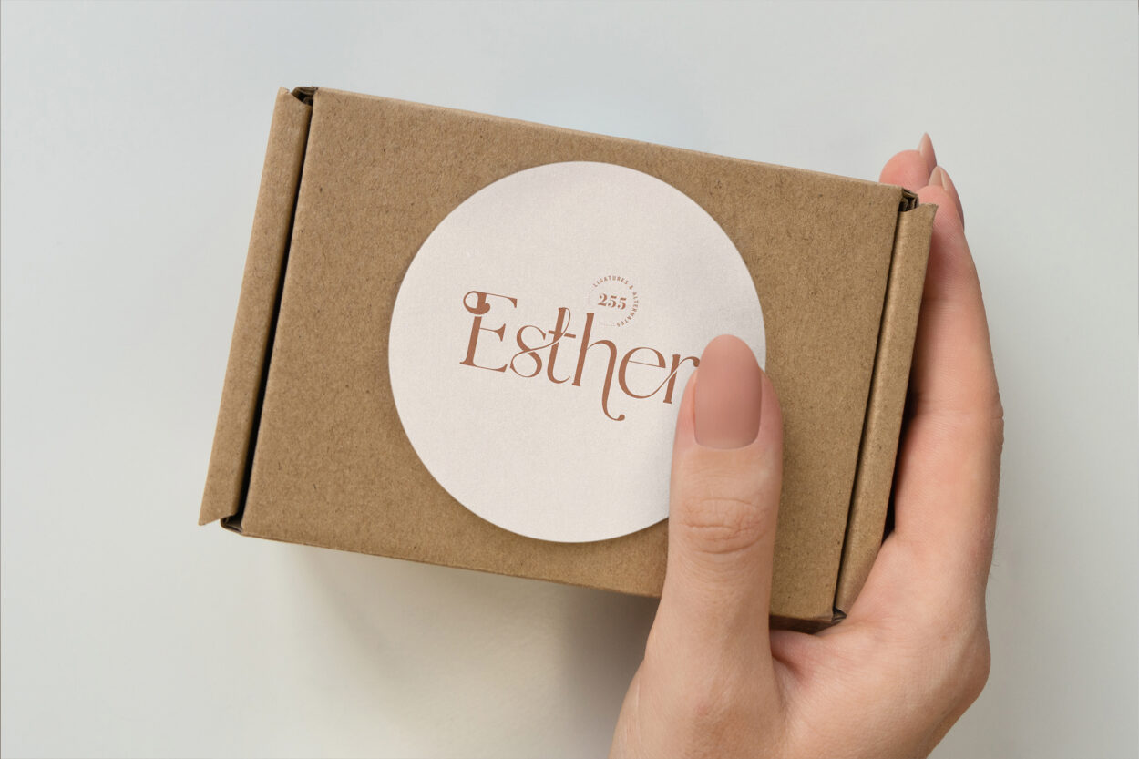 A hand holding a small brown cardboard box with a round white sticker displaying the brand name Esther in elegant typography.