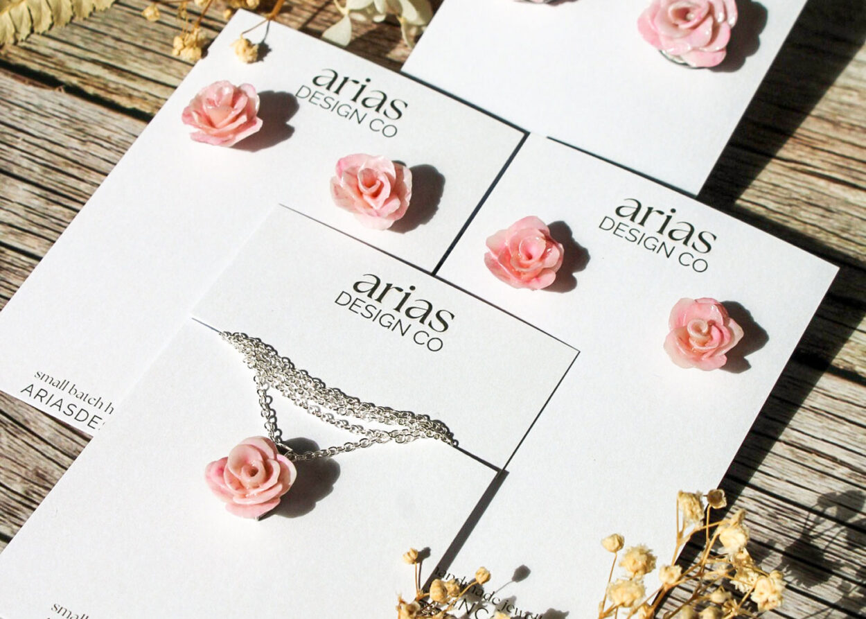 Jewelry display cards from Arias Design Co featuring pink rose earrings and a matching rose pendant necklace on silver chains, placed on a rustic wooden surface with dried flowers as accents.