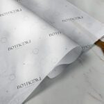 A sheet of white wrapping paper with "Botticelli" branding in black text and subtle decorative elements, partially rolled open on a marble surface with a pair of gold-handled scissors nearby.