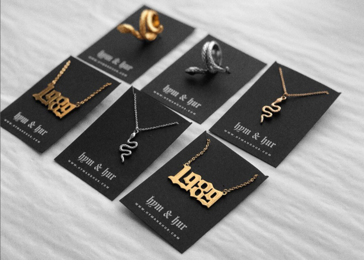 A collection of gold and silver jewelry, including necklaces and rings with unique designs like snakes and text, displayed on black backing cards featuring the 'hym & hur' branding.