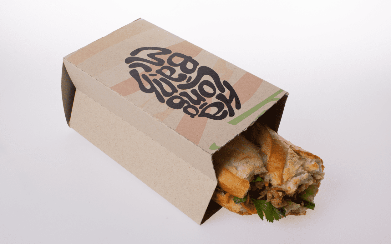 A cardboard sandwich holder with bold black lettering reading 'Ha Long Banh Mi,' partially enclosing a baguette sandwich filled with meat, vegetables, and fresh herbs.