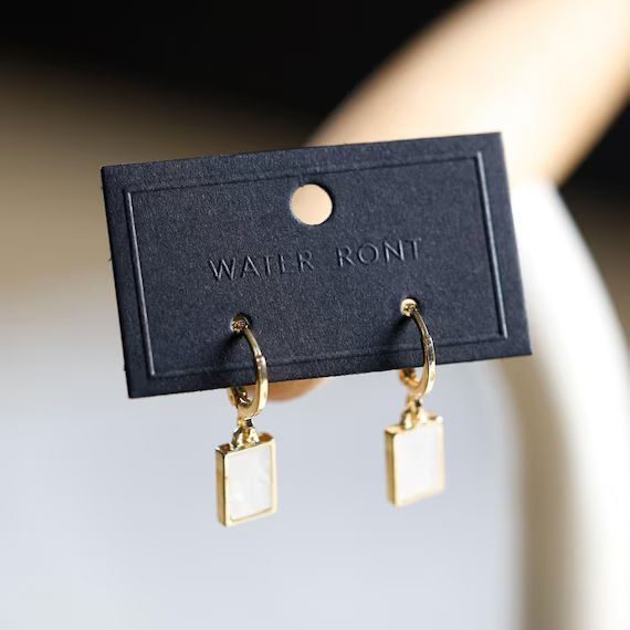 Pair of gold rectangular dangle earrings with a glossy white inset, displayed on a minimal black earring card embossed with the text 'Water Front'.