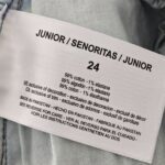 A white clothing label displaying size "24" and the text "Junior / Señoritas / Junior," made of 99% cotton and 1% elastane. The label includes multilingual manufacturing details indicating it was made in Pakistan and care instructions on the reverse side.